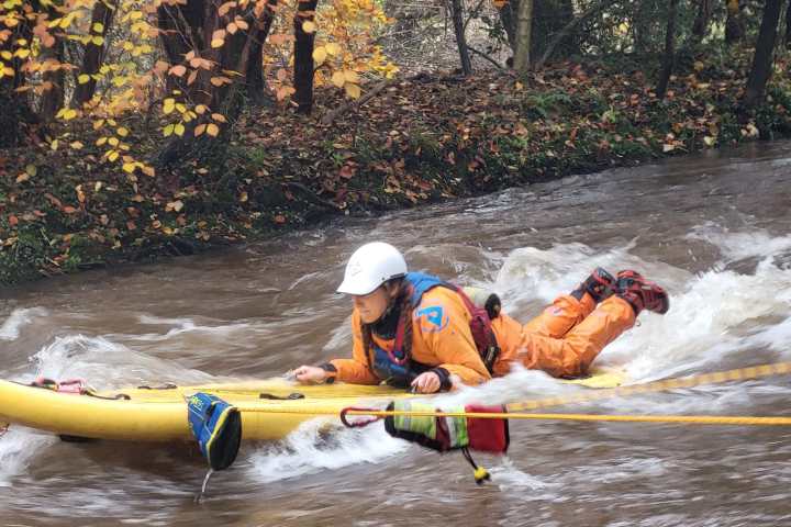 Person in orange suit and helmet on a yellow board in a fast-flowing river, surrounded by autumn leaves.