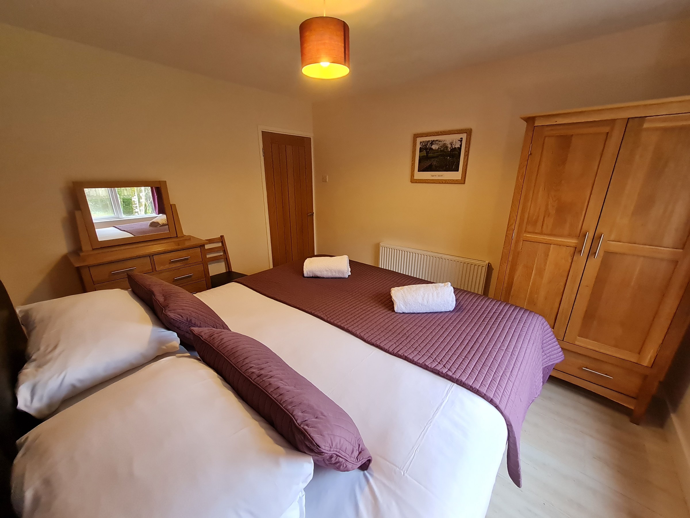 Bedroom with a double bed, purple bedding, wooden wardrobe, dresser, and a ceiling light.