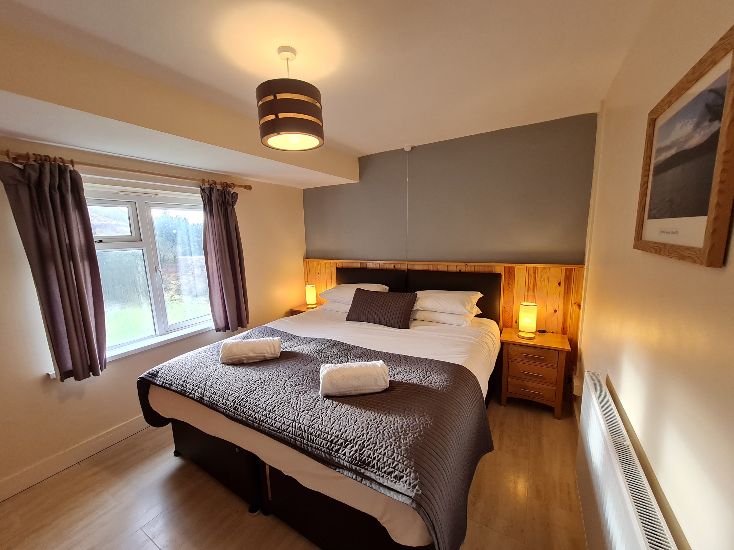 Cozy bedroom with double bed, bedside tables, lamps, window with curtains, and framed picture on wall.
