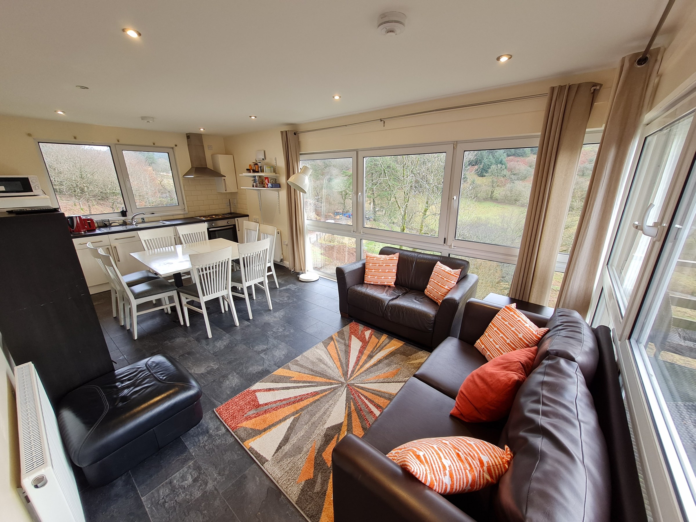 Modern living room and kitchen with sofas, dining table, and large windows.