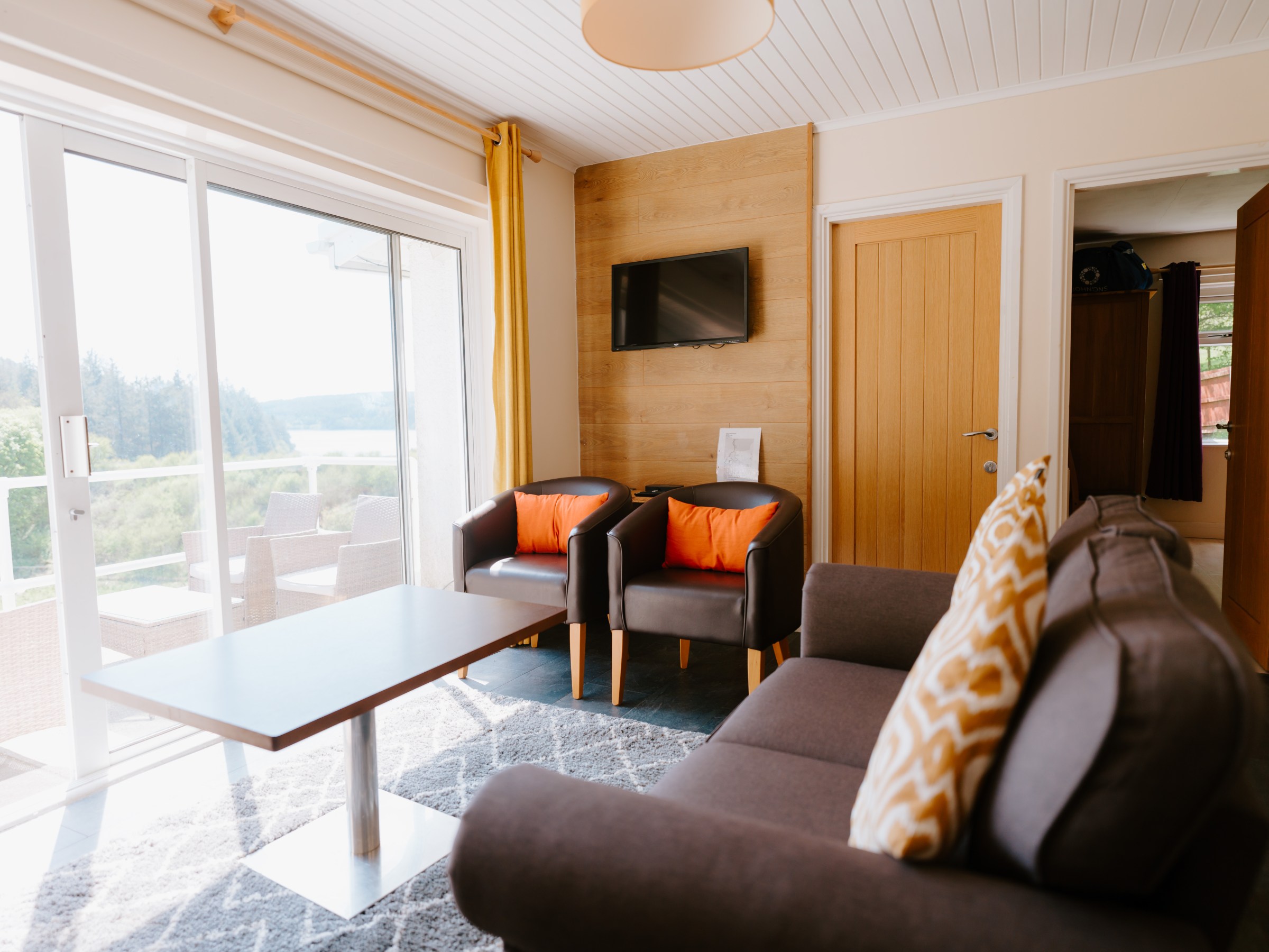 Bright living room with brown sofa, chairs, TV, and glass doors to balcony with outdoor view.