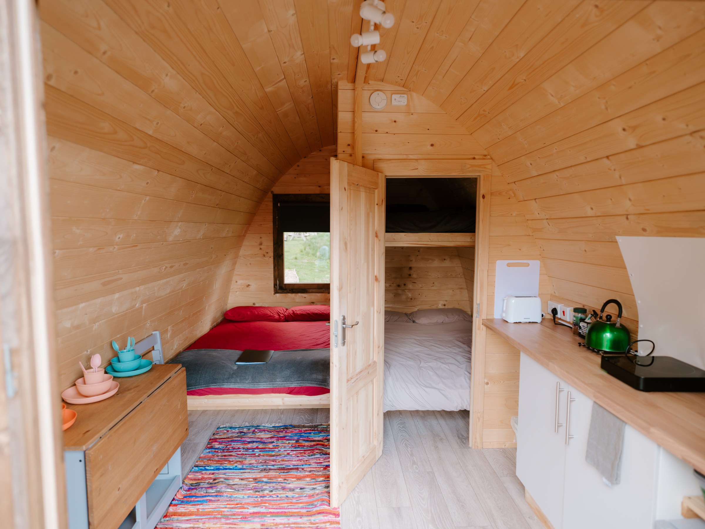 Cozy wooden cabin interior with two beds, kitchenette, and colorful rug.