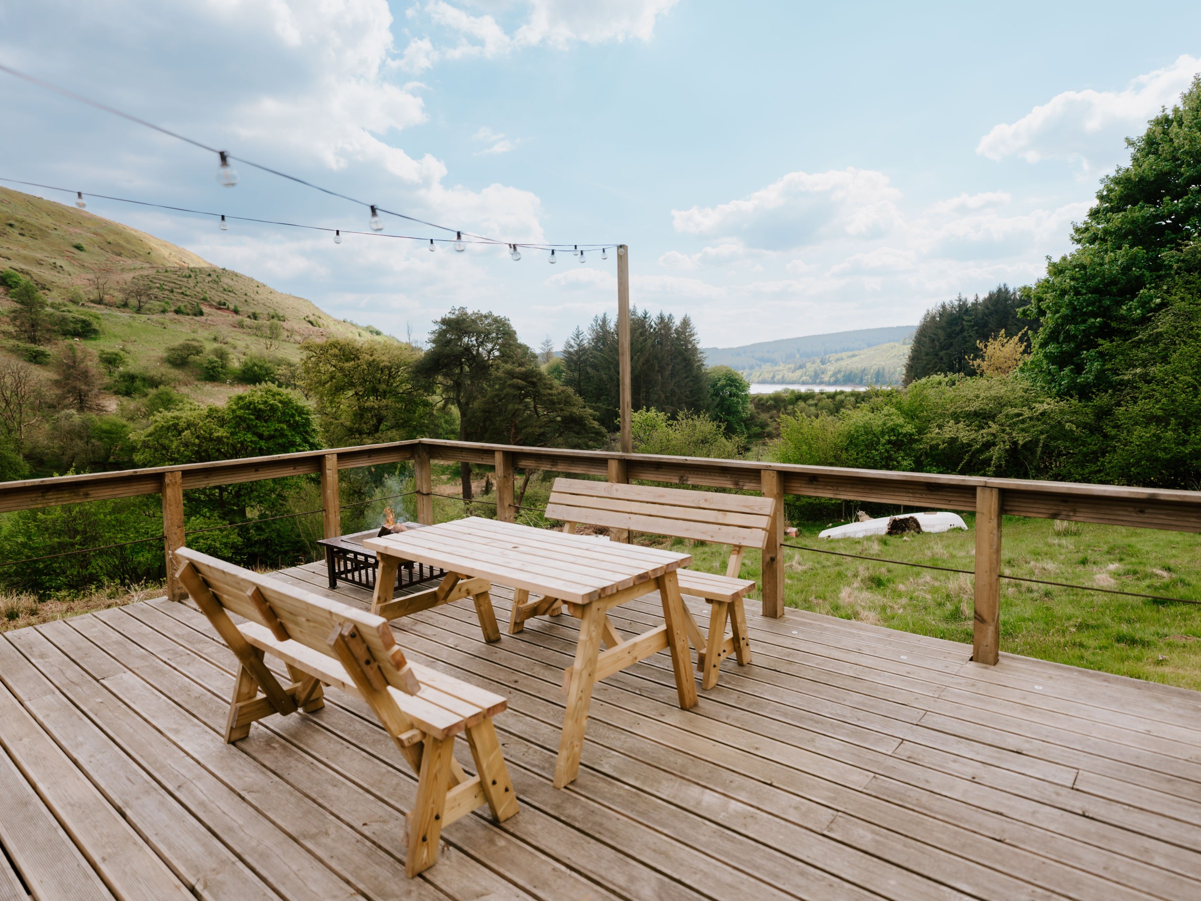 Wooden deck with table, chairs, and scenic view of hills and trees under a blue sky.