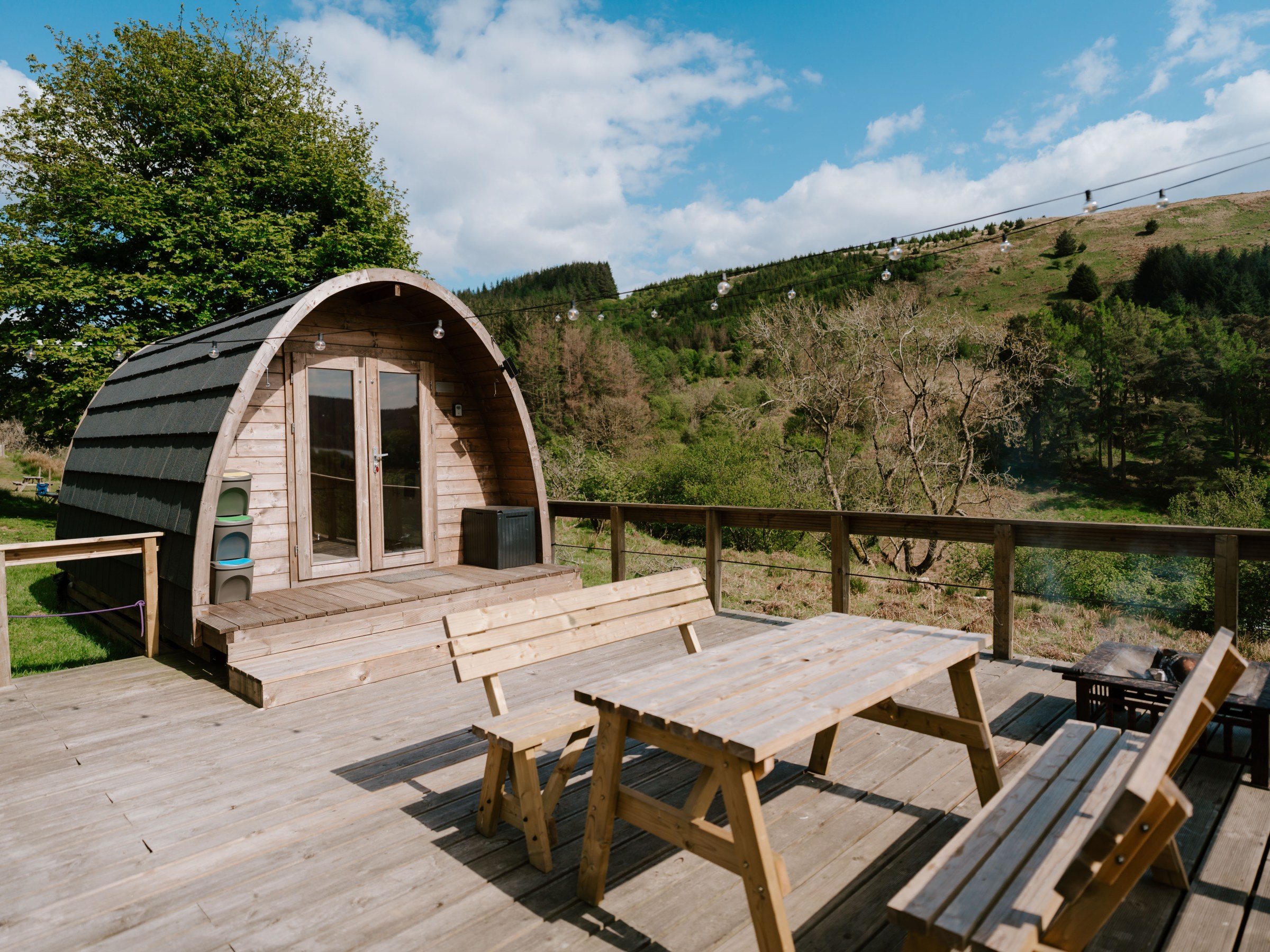 Wooden cabin on a deck with benches, surrounded by trees and hills.