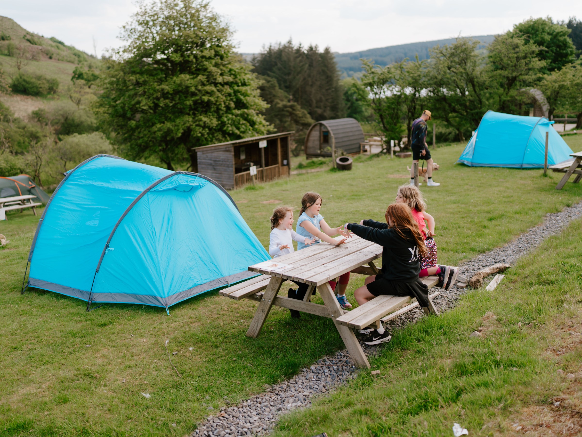Group of young people at picnic table near blue tents in a grassy campsite.
