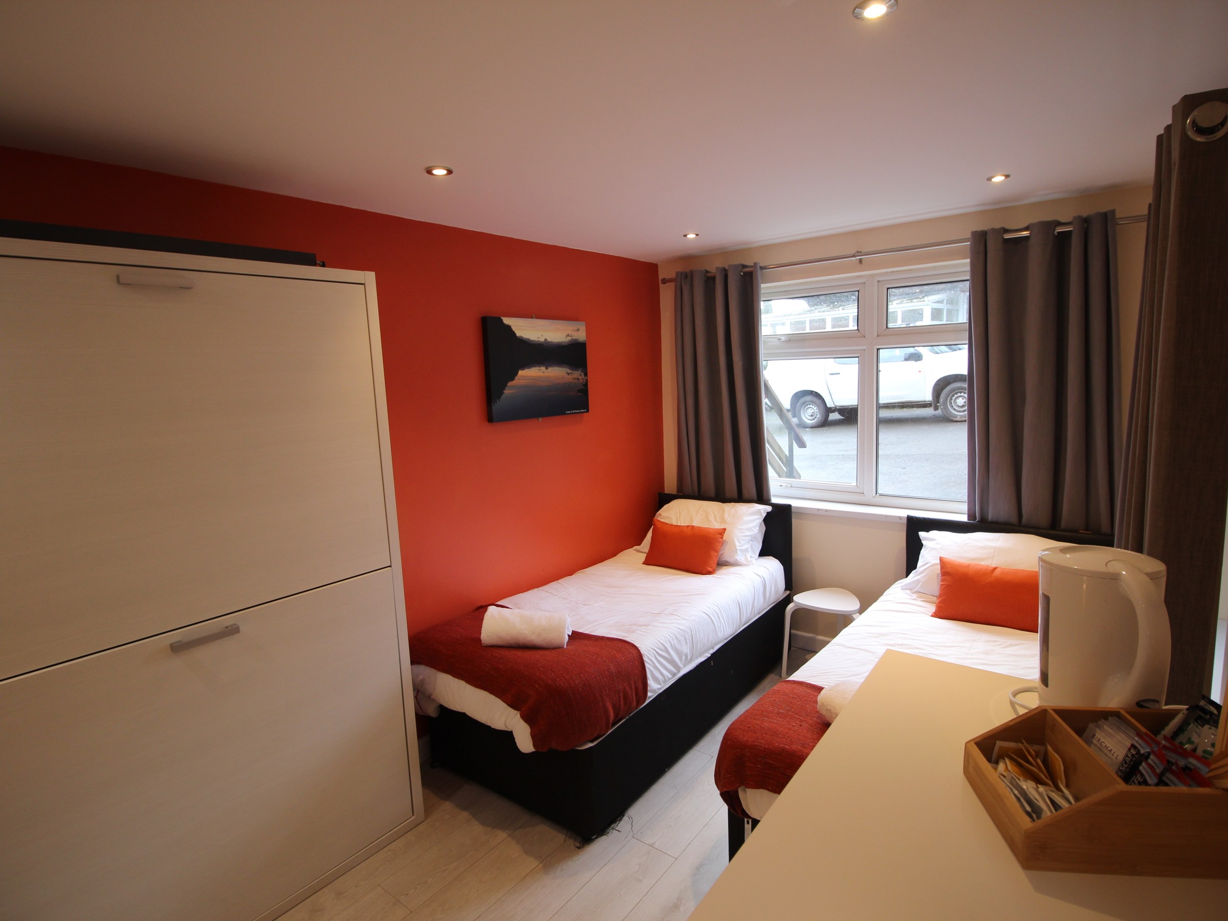 Cozy room with two single beds, orange wall, window view, and small desk.