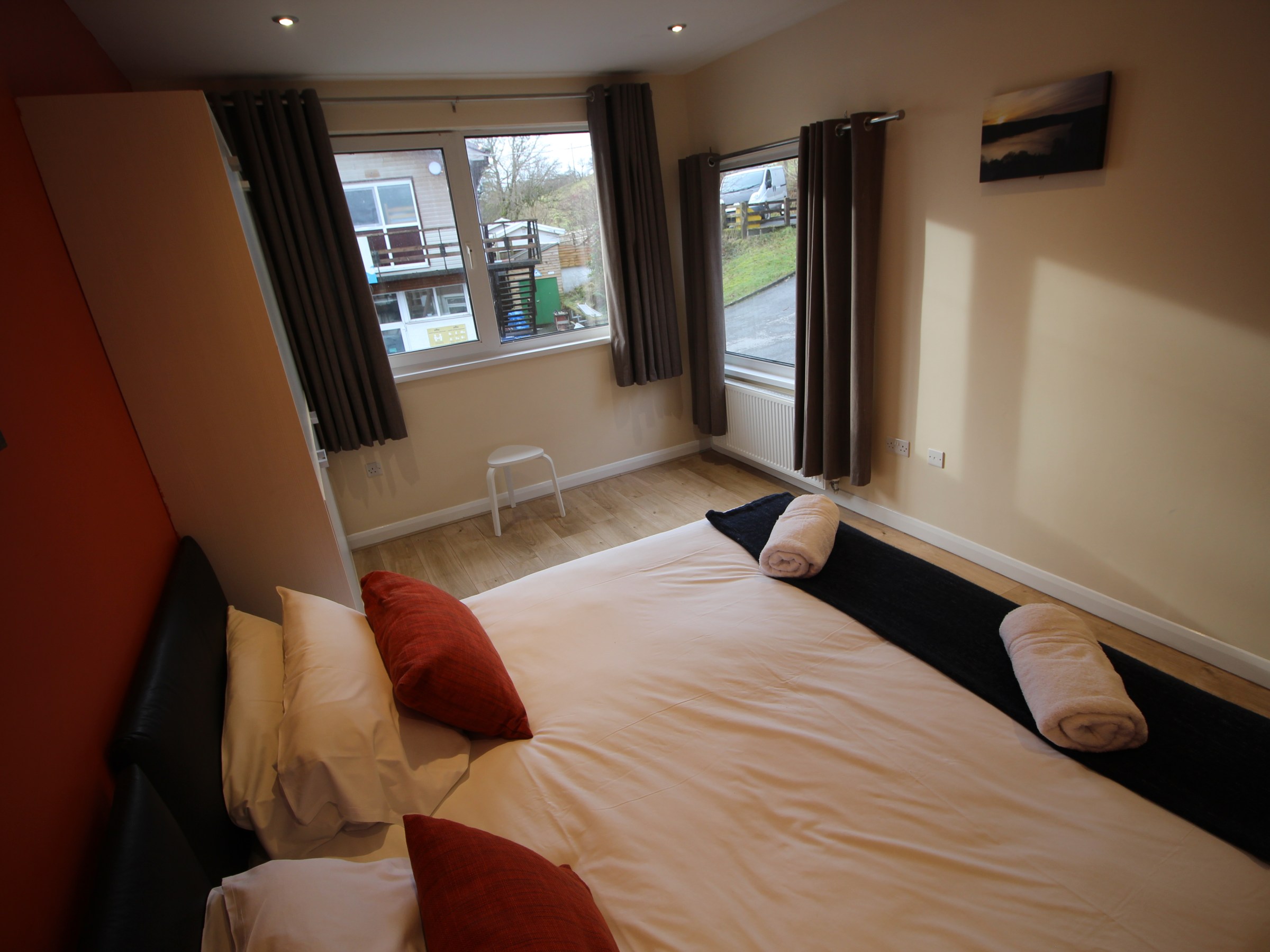 Bedroom with two windows, bed, pillows, and towels on bed, view of houses outside.