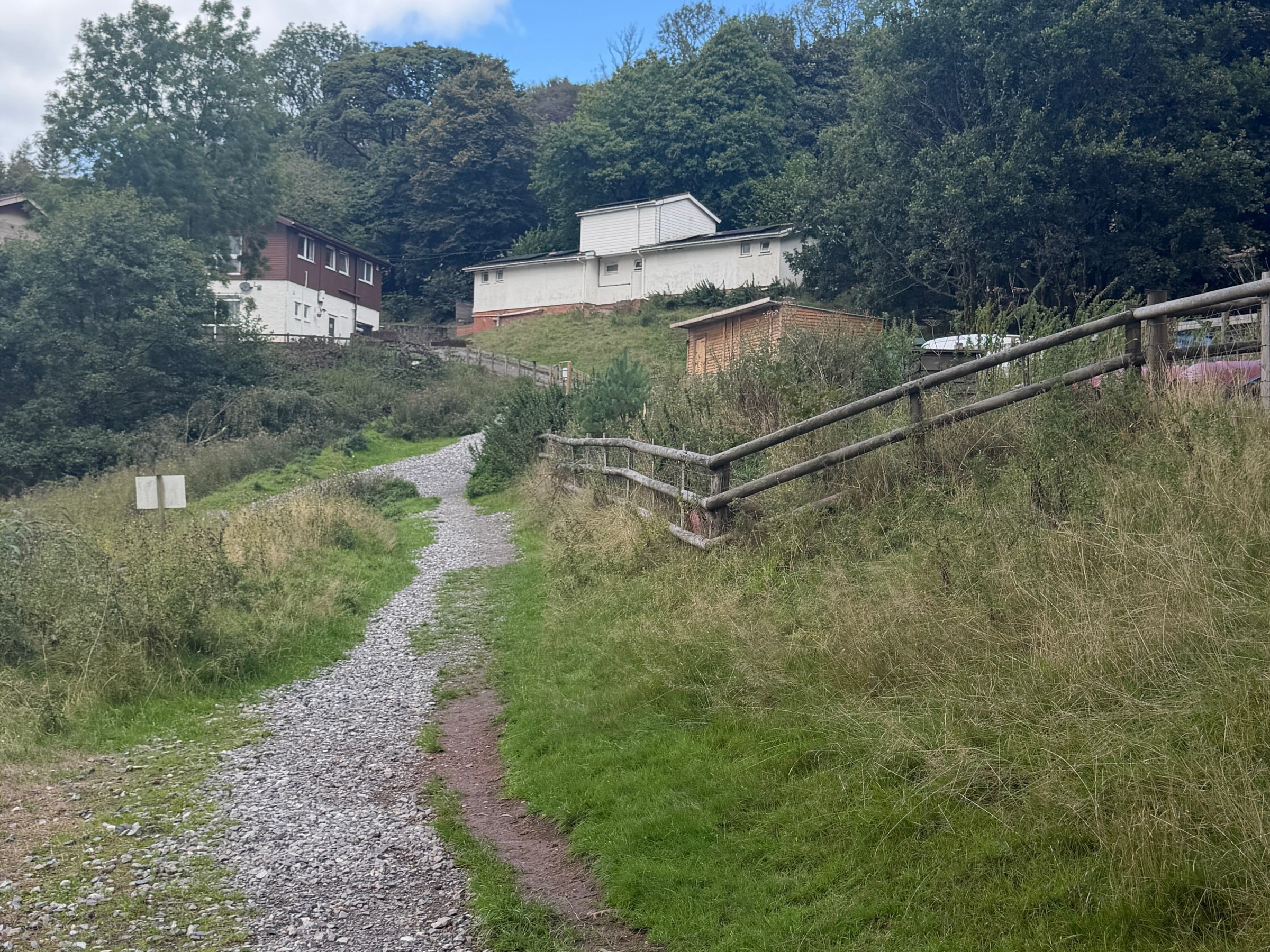 Gravel path leading uphill through grassy area with trees and buildings in the background.